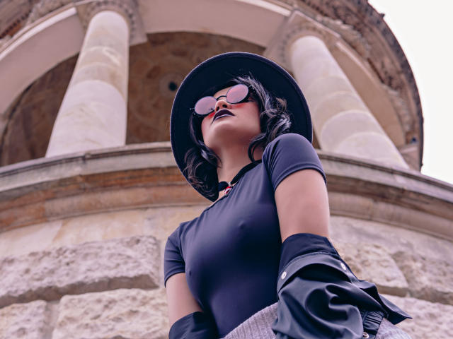 CassieJones with dark wavy hair, black hat, pink sunglasses, black t-shirt, black gloves, and dark lipstick, standing in front of large stone columns, looking down.
