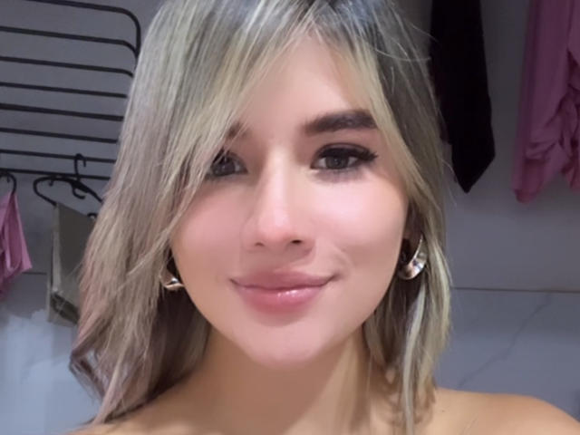SashaPolet with blonde hair, wearing earrings, smiling, showing her pinkish lips, and her small tits are barely visible. She's got a cute face and is looking at the camera.