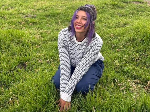 DarckVelvet has purple hair, a white and black patterned sweater, and blue pants, sitting on grass with a cute smile, looking happy and relaxed.
