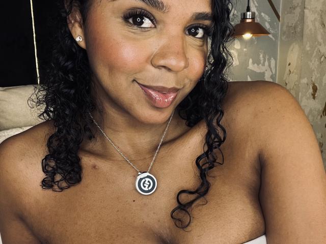LisaMendez has dark curly hair, brown eyes, and a cute smile with glossy pink lips. She's wearing a silver necklace with a spiral design, showing her bare shoulders.