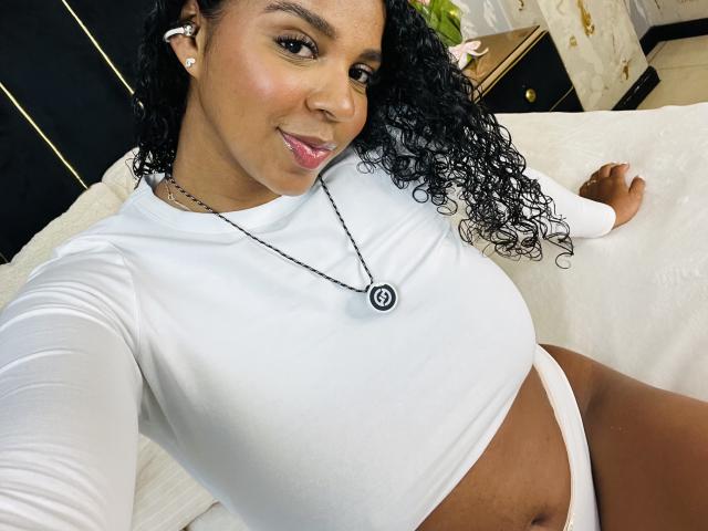 LisaMendez with curly black hair, wearing a white long-sleeve crop top and white panties, showing her tits and a necklace with a black circular emblem. Smiling, sitting on a bed.