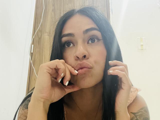 StellaBeckker with long black hair, pouty lips, nose ring, and manicured hands, looking up with a slight pout, in a selfie. Her tits are partially visible.