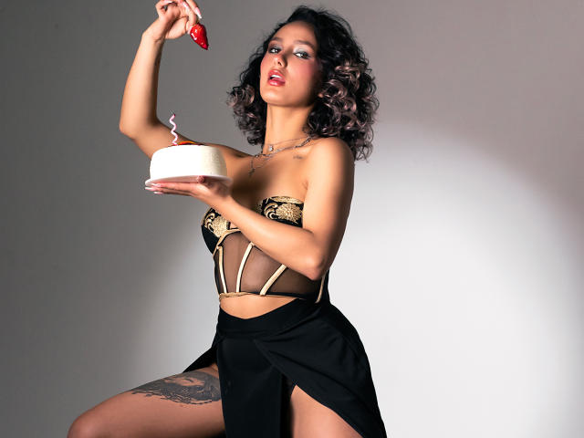 Flora Saenz, met krullend donker haar, houdt een rode aardbei boven haar borsten, en een witte taart met een kaarsje, terwijl ze een zwart en gouden lingerie-set en een zwarte rok draagt..