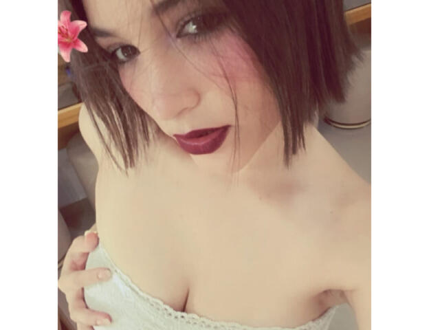 MelanyRios with short brown hair, pink flower in hair, dark lipstick, white towel covering her tits, hand on her chest, looking up with parted red lips.