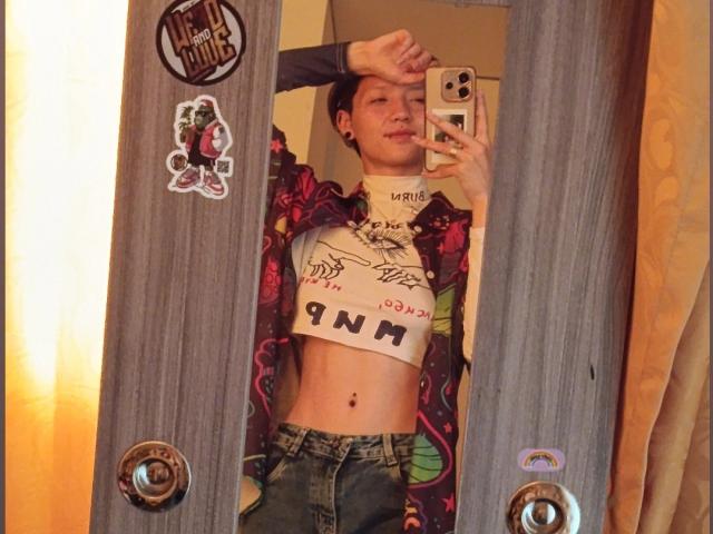 LissyLuxe with short brown hair, wearing a floral jacket, white crop top with "NNN" on it, and blue jeans, taking a mirror selfie, showing her toned midriff.