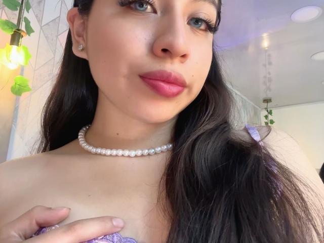 ZoeRedds with long dark hair, pink lipstick, pearl necklace, and purple lace top, showing her small tits with a hand. She's smiling, with green plant leaves in the background.