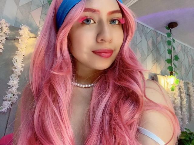 ZoeRedds with pink hair, blue headband, pink eyeliner, pearl necklace, and pink lips, showing off her tits in a cute selfie, wearing a white strap.