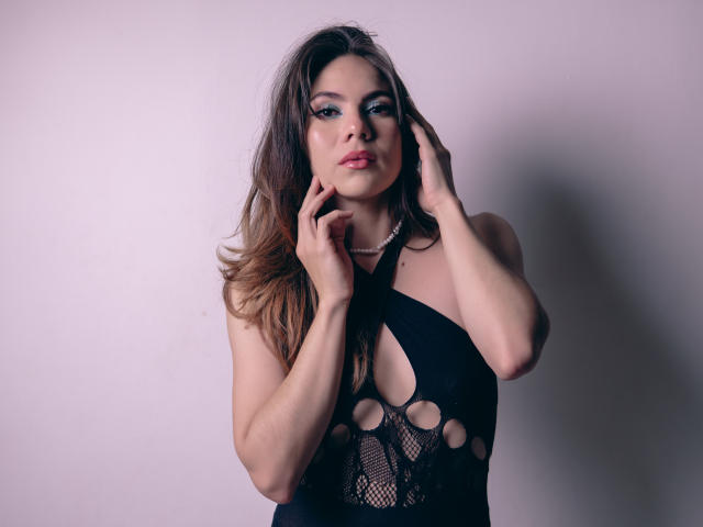 Linda Perez, com cabelos castanhos longos, sombra azul e batom rosa, vestindo um vestido preto com recortes, com as mãos perto do rosto, com uma expressão sedutora, mostrando os seios..
