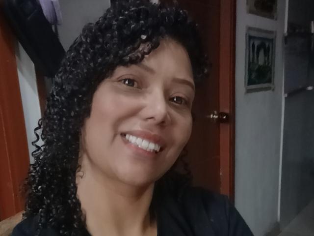 KenyaKarath a les cheveux noirs bouclés, les yeux marron et un sourire qui révèle des dents blanches. Elle a un teint brun clair et porte une chemise noire..