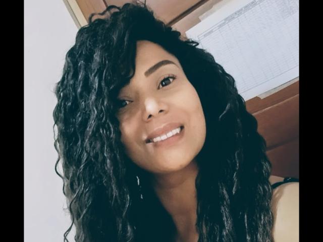 KenyaKarath, con capelli neri ricci, sorride mostrando i denti bianchi e ha gli occhi marroni, indossa una maglietta nera, in un selfie. Le sue natiche non sono visibili..