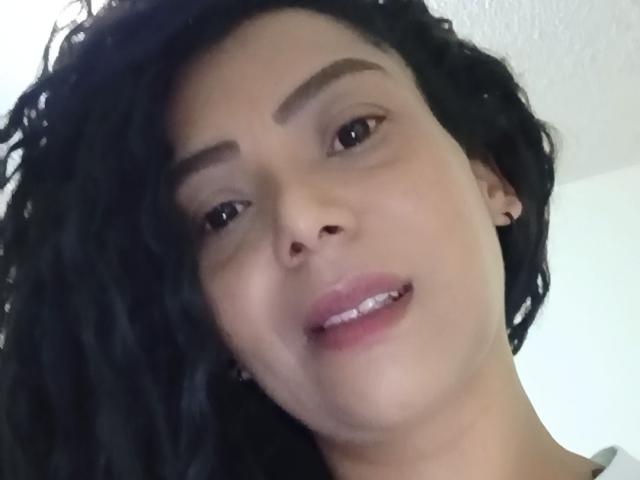KenyaKarath mit lockigem, schwarzem Haar, heller brauner Haut und braunen Augen, die ein leichtes Lächeln mit rosafarbenen Lippen zeigt, trägt kleine Ohrringe und blickt leicht nach rechts..