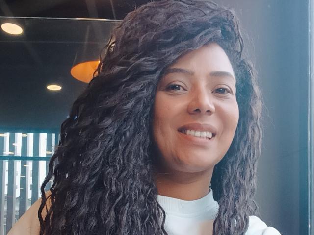 KenyaKarath ha i capelli neri ricci, un sorriso carino e la pelle marrone chiaro. Indossa una maglietta bianca e la luce del sole mette in risalto la sua espressione felice..