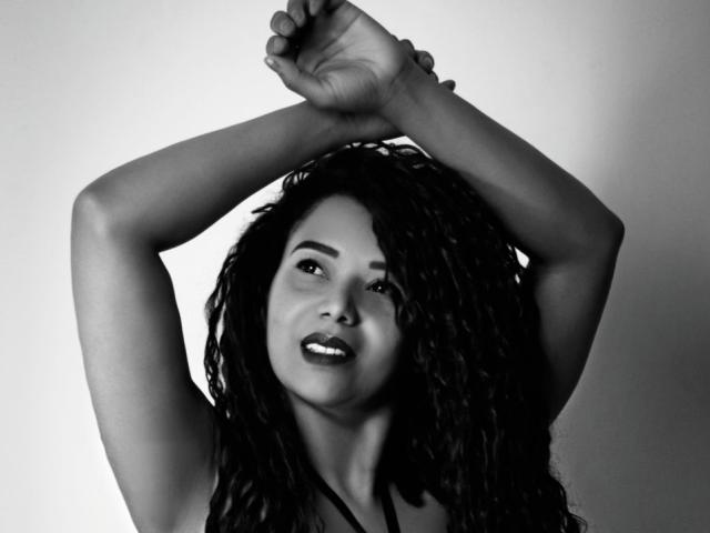 KenyaKarath, con le braccia incrociate, fotografia in bianco e nero, capelli ricci scuri, sorride leggermente, carnagione media, indossa un top nero a spalline, guarda di lato, sexy..