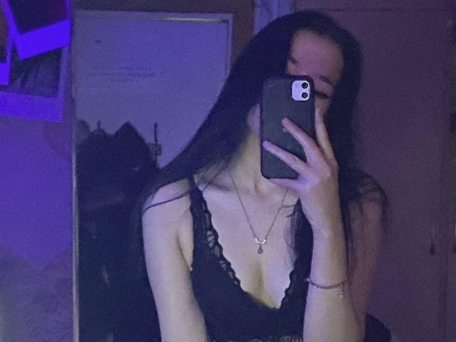 MertieDasinger, con lunghi capelli neri, indossa un reggiseno di pizzo nero, tiene un telefono, si copre il viso, mostrando il seno, in una stanza poco illuminata, e indossa una collana..
