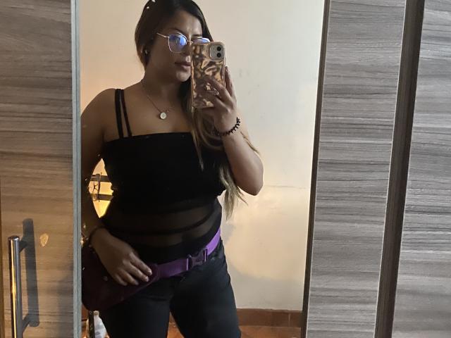 TompsonKarina with glasses, black top, and purple belt, showing her tits in a mirror selfie, long brown hair, bracelet, and phone with a patterned case.