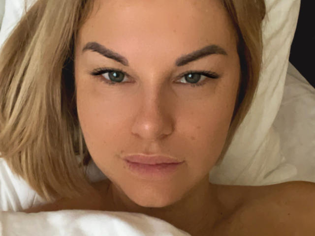 BelleLisaG's face, lying in bed, with blonde hair, green eyes, and a slight smirk, showing her bare shoulders. White pillow and sheets in the background.
