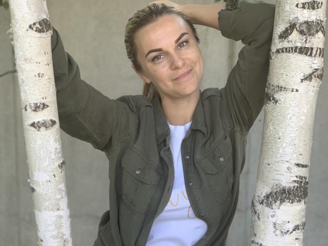 BelleLisaG with arms raised, holding birch trees, wearing an open green shirt, white tee, smiling, blonde hair tied back, blue eyes, fair skin, standing outdoors, casual look.