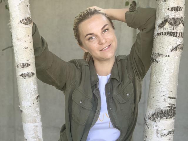 BelleLisaG with blonde hair in a ponytail, green shirt, white tee, arms up holding birch trees, smiling, freckles, blue eyes, slight tan, relaxed, natural light.