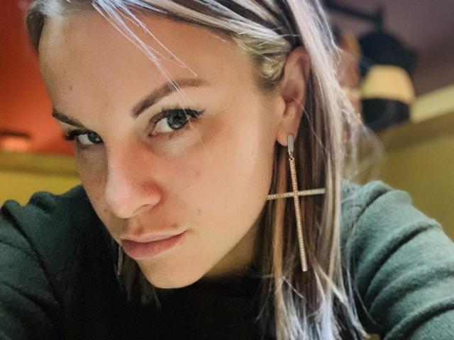 BelleLisaG with blonde hair, green eyes, wearing a cross earring, and a green hoodie, has a slight pout, looking directly at the camera. Her face is centered.