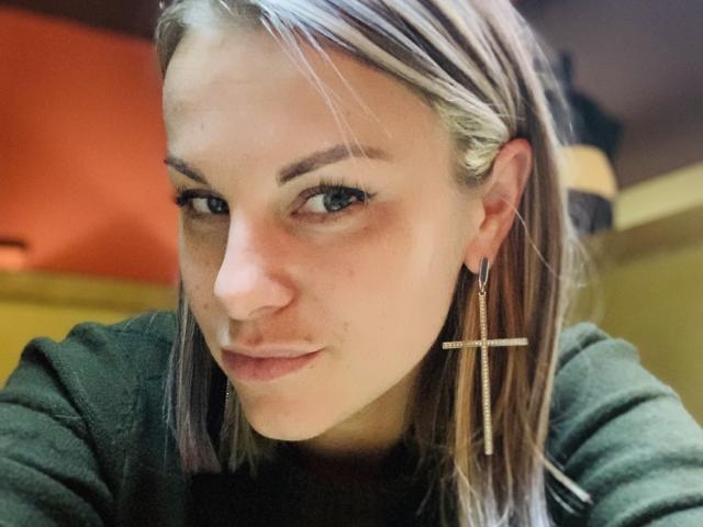 BelleLisaG has pale skin, blonde hair with braids, green eyes, and a small cross earring in her left ear. She's wearing a dark green shirt, looking at the camera.