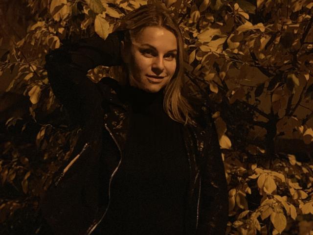 BelleLisaG mit blonden Haaren, schwarzem Rollkragenpullover und schwarzer Jacke, Hand im Haar, lächelnd, umgeben von braunen, gelben Blättern, gedämpftes, warmes Licht, leicht zur Seite blickend..