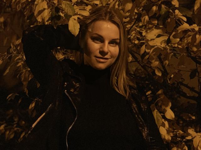 BelleLisaG mit blonden Haaren, dunklem Rollkragenpullover, schwarzer Jacke, Hand im Haar, lächelnd, schwach beleuchtet, warme braune Töne, Blätter im Hintergrund, weiches Licht, sexy, selbstbewusst, attraktiv..