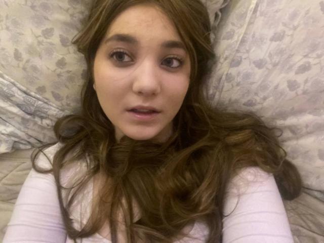 MichikoVivona has long brown hair, a cute face, and is looking up with brown eyes, wearing a white shirt, lying on a patterned pillow, with her mouth slightly open.