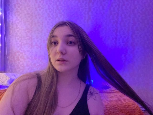 MichikoVivona with long brown hair, wearing a black tank top, has a small nose piercing and a tattoo on her right shoulder, looking slightly bored under blue lighting.