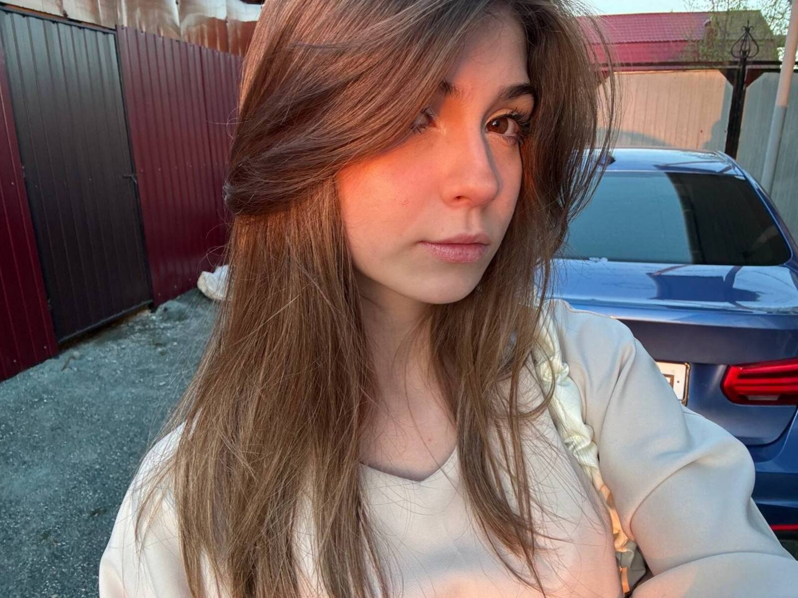 LauraleePenderel with long brown hair, white shirt, and serious expression, standing in front of a blue car, sunlight casting a warm glow on her face and chest.