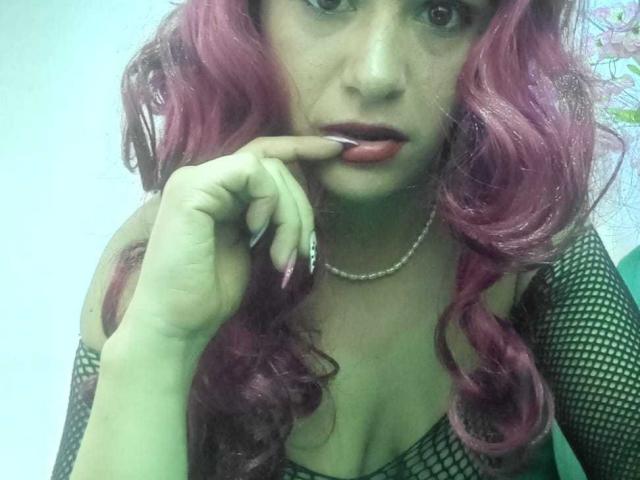 AriannaSeete with pink wavy hair, green fishnet top, and pearl necklace, biting her red-lipsticked finger, showing off her cleavage. She has a serious, sexy look.