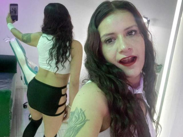 AriannaSeete with long brown wavy hair, red lipstick, white crop top, black strappy shorts, and white thigh-high socks, taking a mirror selfie, showing her tattooed arm and ass.