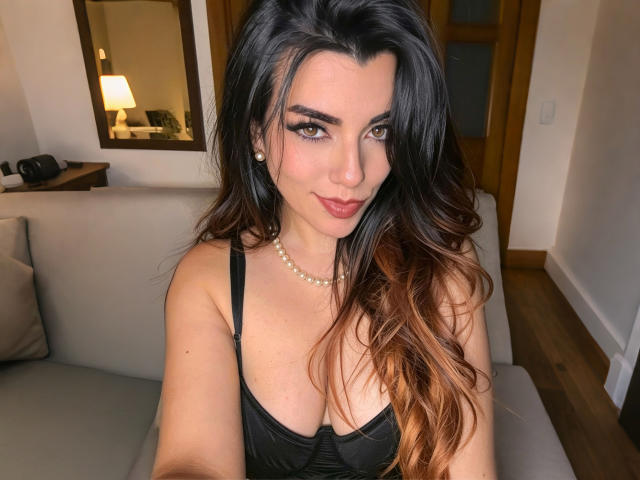 ZoeeMar ha lunghi capelli castano scuro ondulati, occhi marroni, e indossa un vestito nero con una collana di perle, seduta su un divano grigio, mostrando il suo seno..