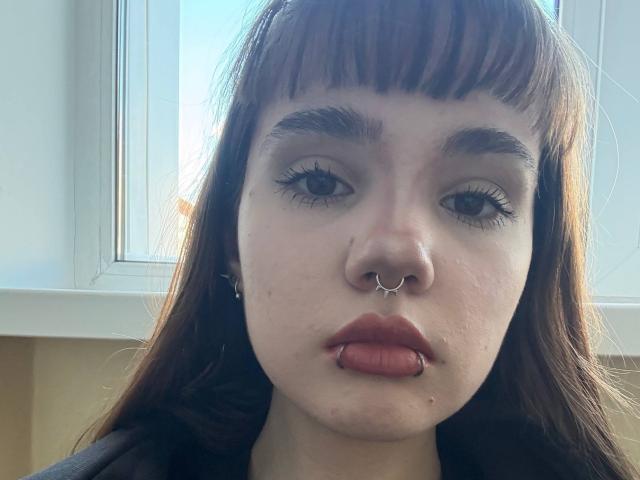 ElseEikner has pale skin, brown bangs, a nose ring, and a lip ring. Her brown eyes have long lashes, and she's looking straight at the camera.