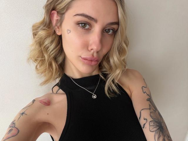 SensualMila with wavy blonde hair, green eyes, and a small heart tattoo on her cheek. She's wearing a black sleeveless top, showing tattoos on her arms. Soft pout.