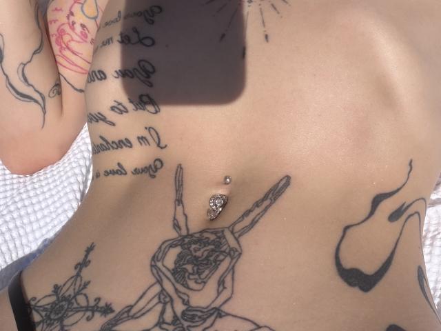 SensualMila's bare, tattooed torso with a belly button piercing and multiple tattoos, including a rose and script. She has light skin and is wearing black underwear.
