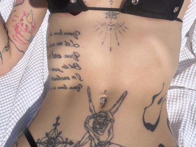 SensualMila's torso, featuring tattoos including a pierced belly button, a smiley face above, a cupcake on the arm, and a rabbit holding a flower, wearing a black bra.