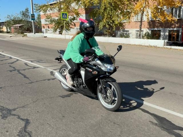 Linette Savel op een zwarte motorfiets, gekleed in een groen shirt, een zwarte helm met roze veren, en zwarte broek, rijdend op een zonnige straat. Haar billen zijn lichtelijk omhoog..