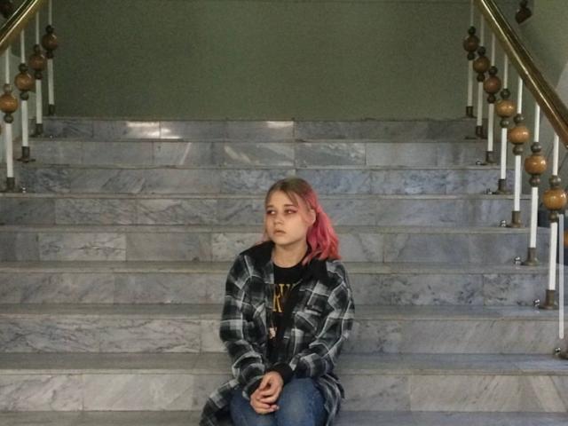 Linette Savel, con i capelli tinti di rosa, è seduta sui gradini di marmo, indossa una giacca a quadri nera e grigia, con le mani sulle ginocchia, e guarda di lato con un'espressione seria..