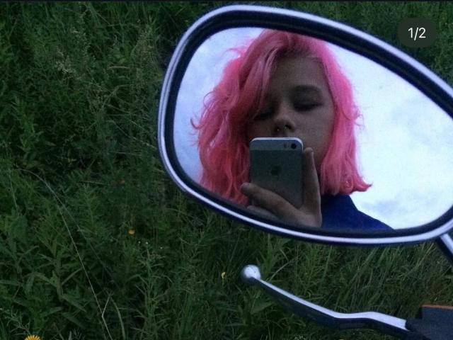 LinetteSavel, con cabello rosa, ojos cerrados, tomando una selfie con un teléfono inteligente en un espejo, reflejada en un espejo lateral, sosteniendo el teléfono con la mano izquierda, con césped en el fondo..