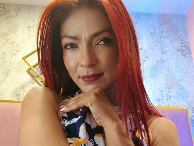 HannaPretty with red hair, wearing a floral top, has a cute smile, dark eyes, and pink lipstick. Her hand rests on her chin, showing off her white nails.