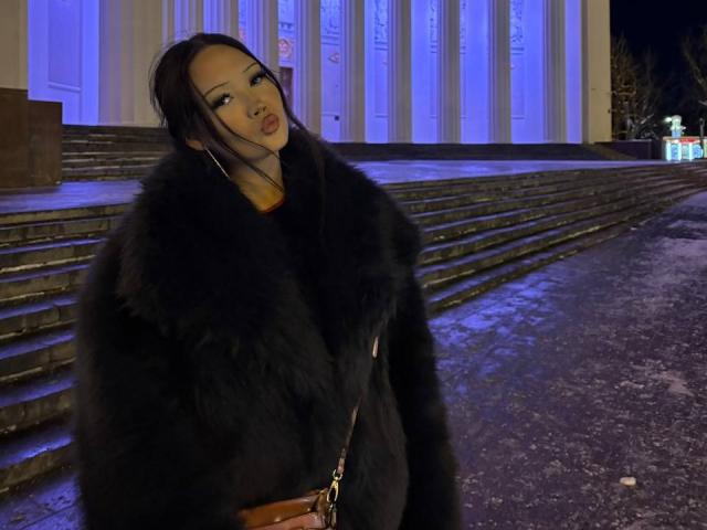 LakendraEytcheson in a black fur coat, standing at night, with blue-lit building steps behind. She has long black hair, heavy makeup, and a brown purse. Snow on ground.