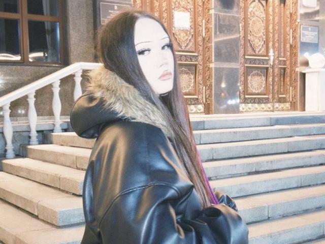 LakendraEytcheson with long brown hair, wearing a black leather jacket with fur trim, standing in front of ornate wooden doors, looking over her shoulder, cold outside.