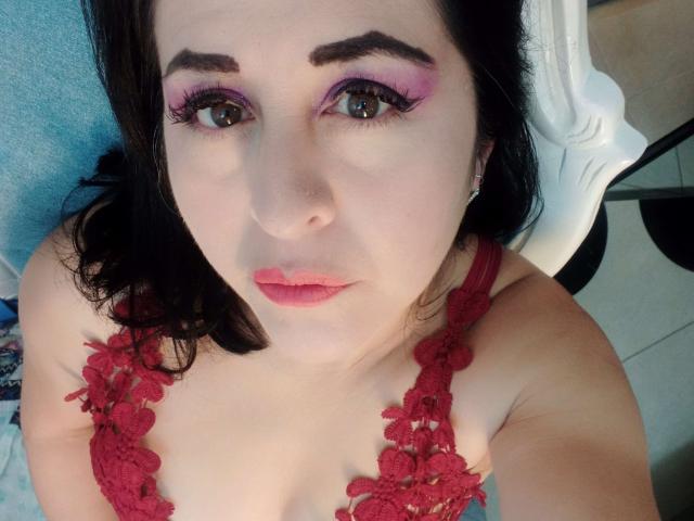 ScarletMilfX com sombra rosa, sutiã de renda vermelha e batom rosa, olhando para cima com olhos castanhos e um leve franzido. Seus seios estão visíveis, e ela tem cabelo escuro..