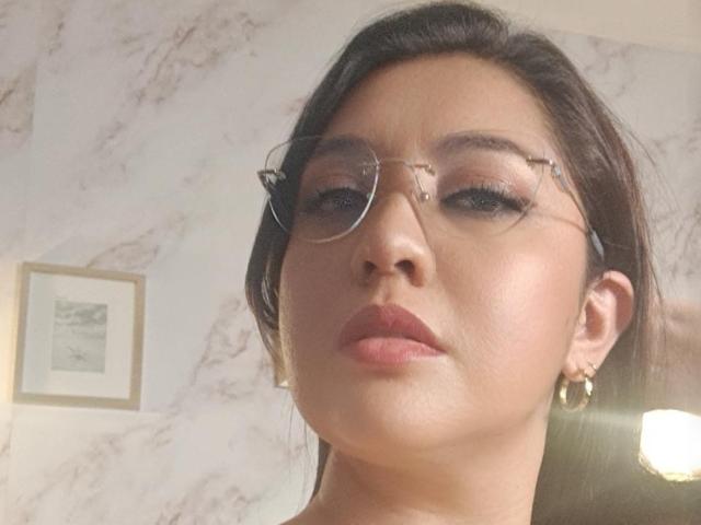 PaulaAnd has glasses, brown hair, and is wearing pink lipstick. Her face shows a neutral expression, and she's wearing gold hoop earrings. Her skin is fair.