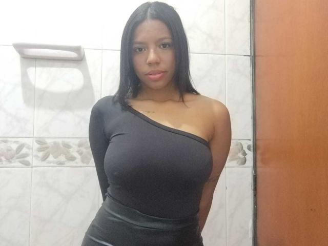 SammyRosi has large, round tits, a small, flat ass, and is wearing a black, one-shoulder, tight dress, with straight black hair, and a neutral expression in a tiled room.