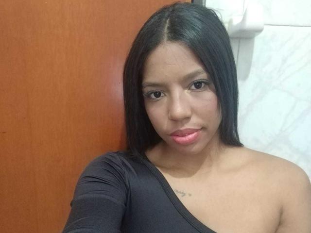 SammyRosi has long black hair, a slight tan, and wears a black, off-shoulder top, showing small tits and a small tattoo near her collarbone, with pink lips.