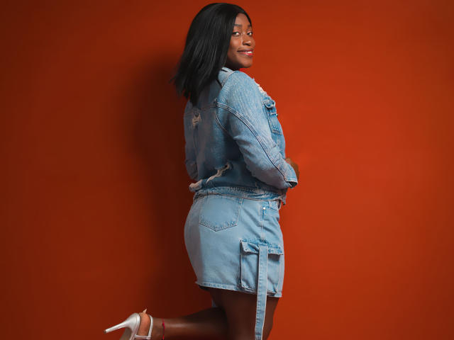 NayaKarter in a denim jacket and skirt, showing her ass, with black hair, smiling, and wearing white high heels, standing against an orange wall.
