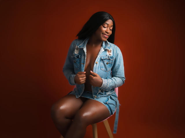 NayaKarter with dark skin, black hair, smiling, wearing a blue denim jacket, showing tits, sitting on a wooden stool, against a red background, hands on jacket.