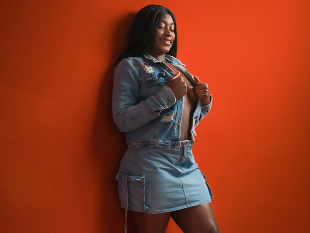 NayaKarter with dark skin, long black hair, and a smile, wearing a ripped blue denim jacket and skirt, exposing her tits, standing against an orange wall.