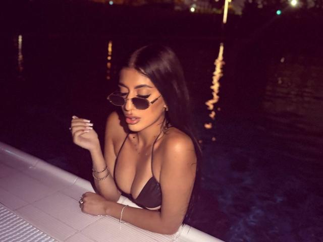 DariaVeil, com longos cabelos pretos, vestindo um minúsculo biquíni preto, mostrando seus seios, em uma piscina escura à noite, usando óculos de sol estilo aviador e segurando um cigarro..